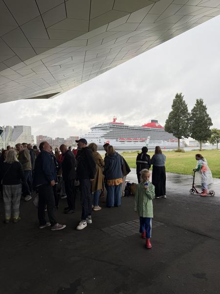 The photo captures a bustling scene at IJpromenade, Overhoeks, Amsterdam, with a large crowd gathered under a modern architectural overhang